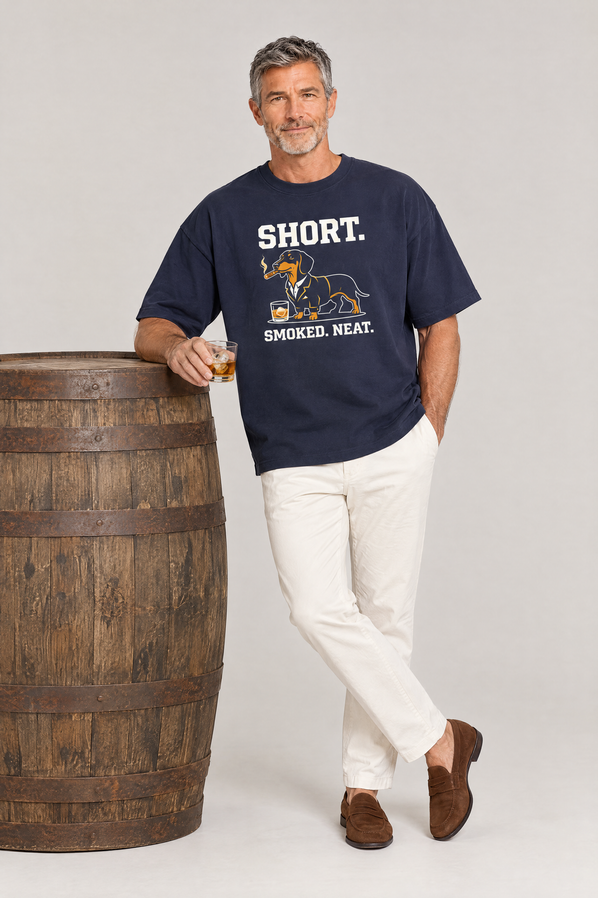 Man leaning on a whisky barrel with a glass in the navy Short Smoked Neat Dachshund Tee. Color: Navy