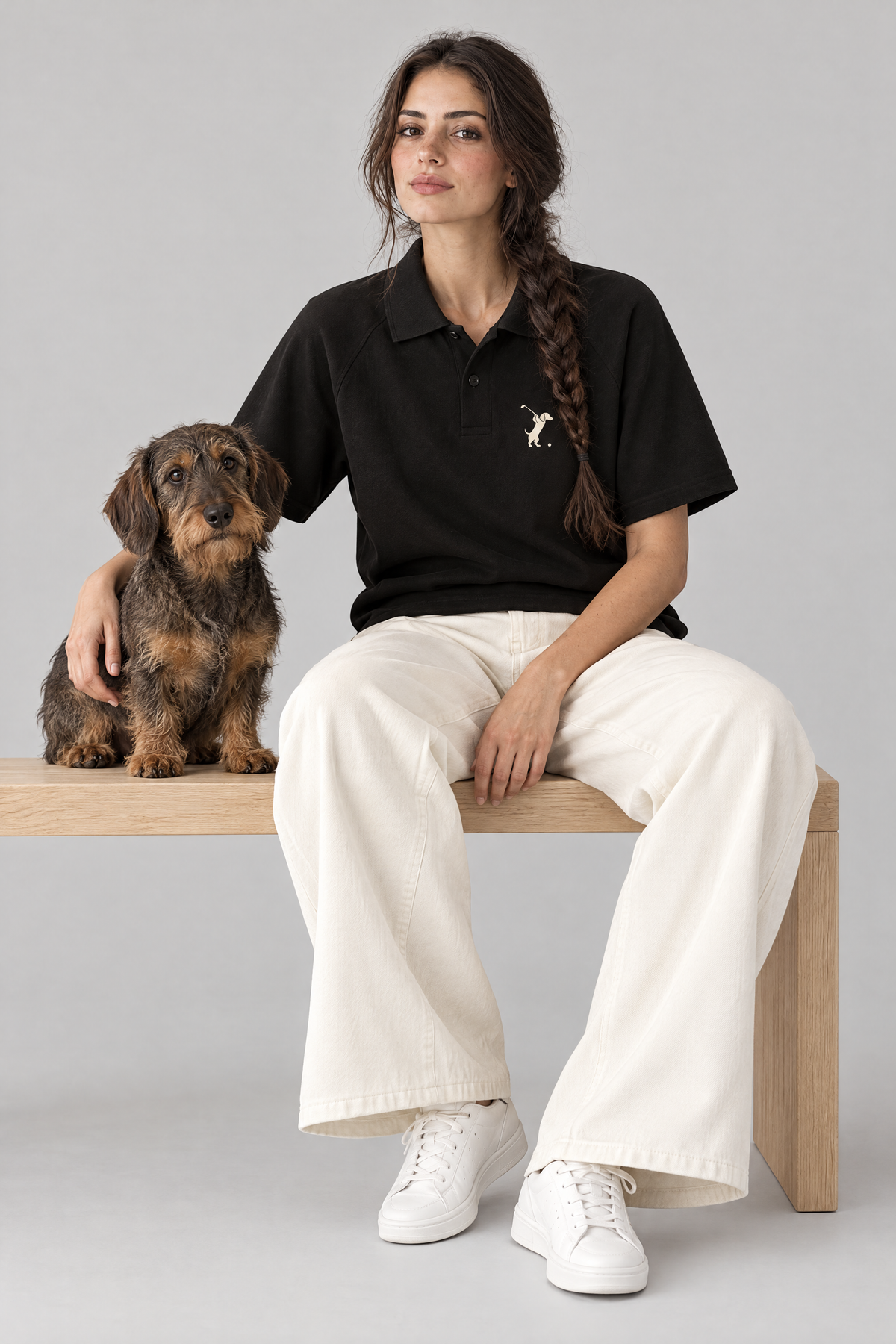 Woman sitting on a wooden bench with a wire-haired dachshund in the black Swing dachshund golf polo and cream wide-leg trousers. Color: Black