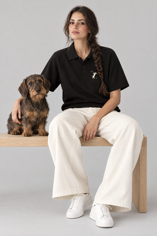Woman sitting on a wooden bench with a wire-haired dachshund in the black Swing dachshund golf polo and cream wide-leg trousers. Color: Black