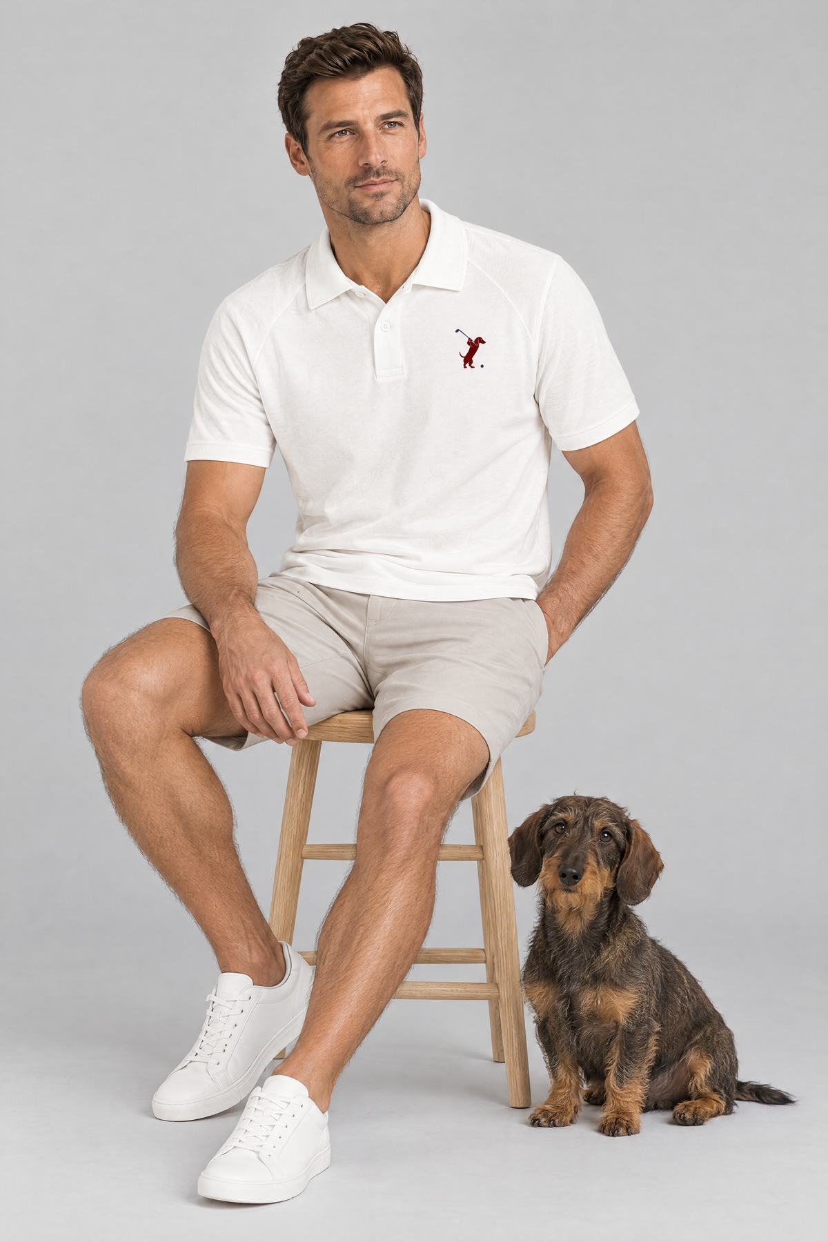 Man sitting on a wooden stool with a wire-haired dachshund in the white Swing dachshund golf polo and stone golf shorts. Color: White