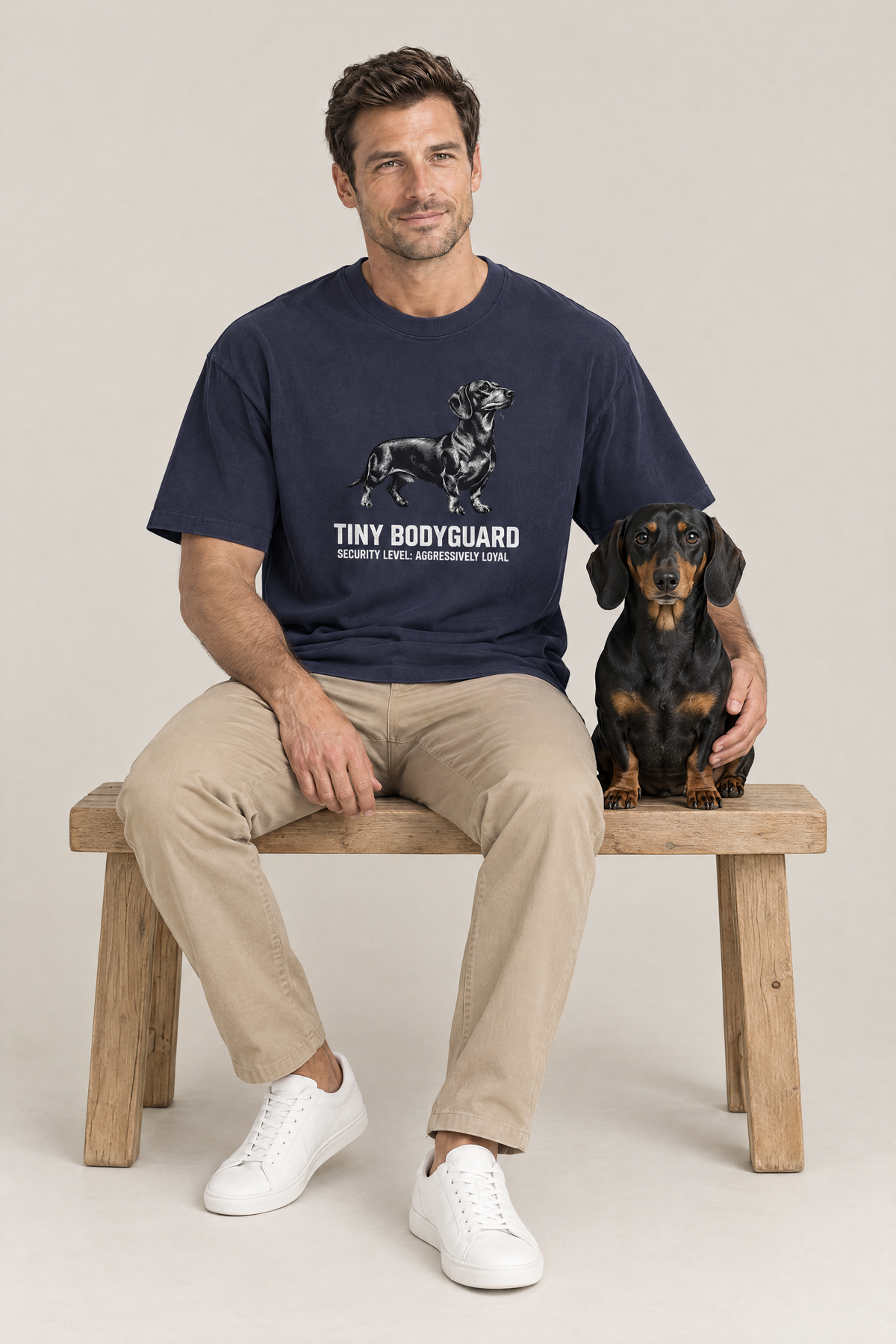Man sitting on a wooden bench next to a black and tan dachshund in the navy Tiny Bodyguard Dachshund Tee. Color: Navy