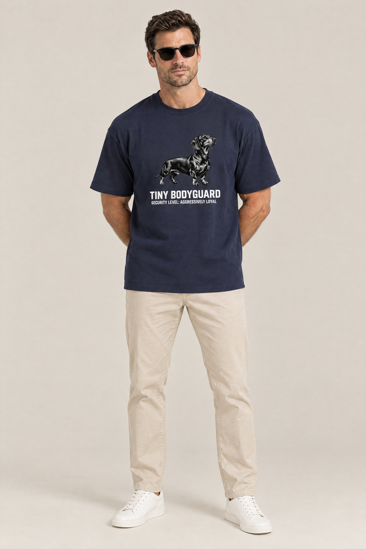 Man in sunglasses wearing the Tiny Bodyguard Dachshund Tee in navy with beige chinos. Color: Navy