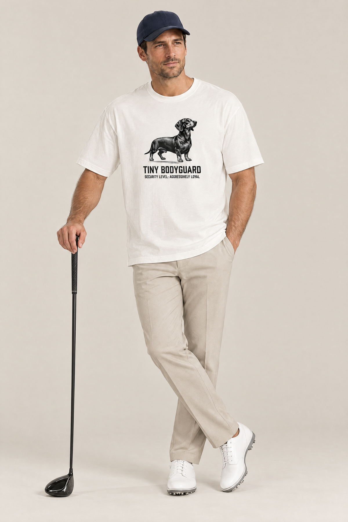Man in a navy cap leaning on a golf driver in the white Tiny Bodyguard Dachshund Tee. Color: White
