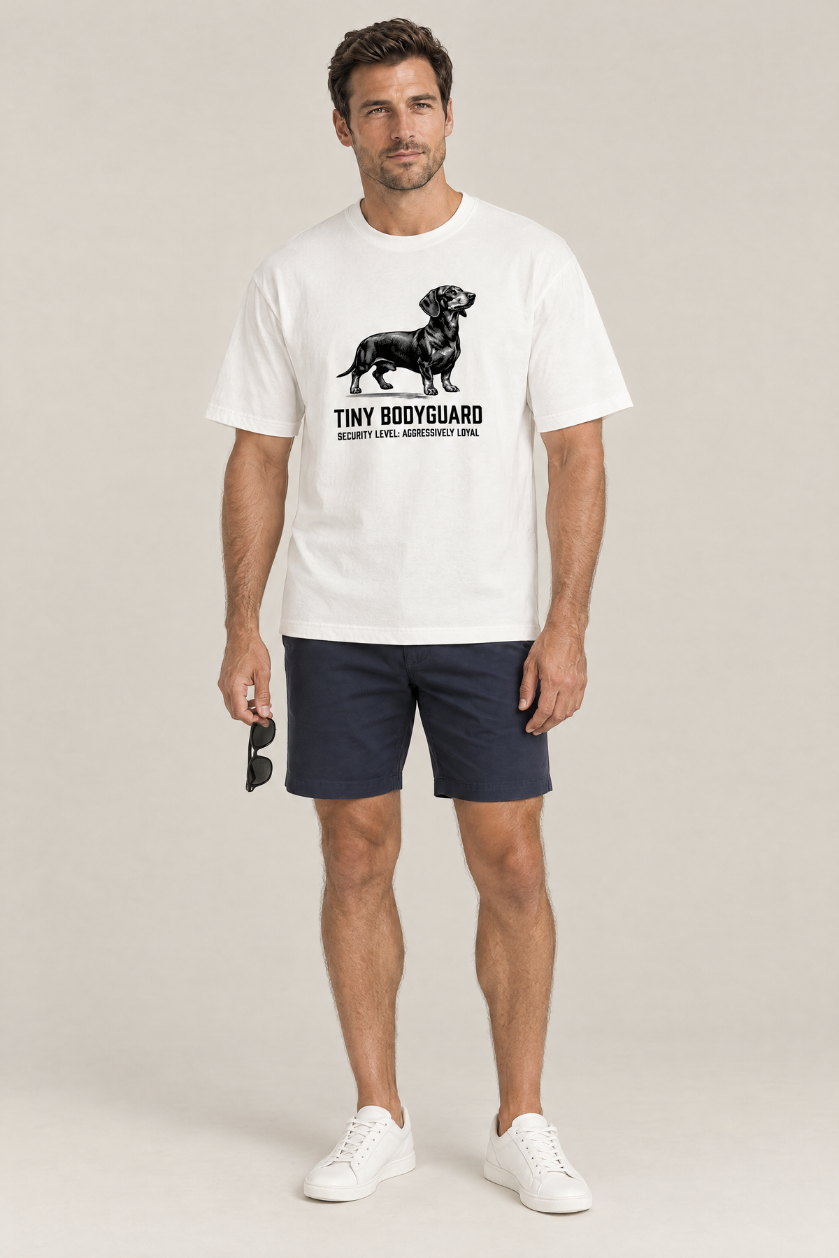 Man wearing the Tiny Bodyguard Dachshund Tee in white with navy shorts and white sneakers. Color: White