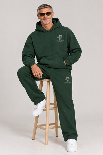 Man seated in The Long Drive dachshund golf hoodie and sweatpants in Blackish Green. Color: Blackish Green