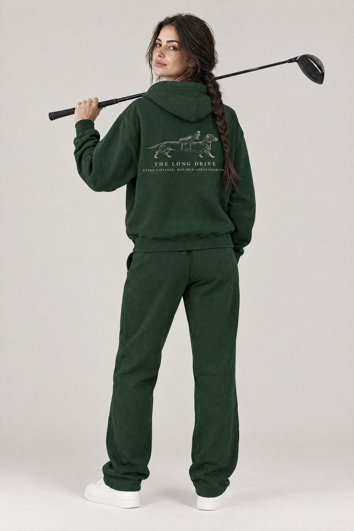 Woman with a driver over her shoulder, back view of The Long Drive dachshund golf hoodie in Blackish Green with the large back print. Color: Blackish Green