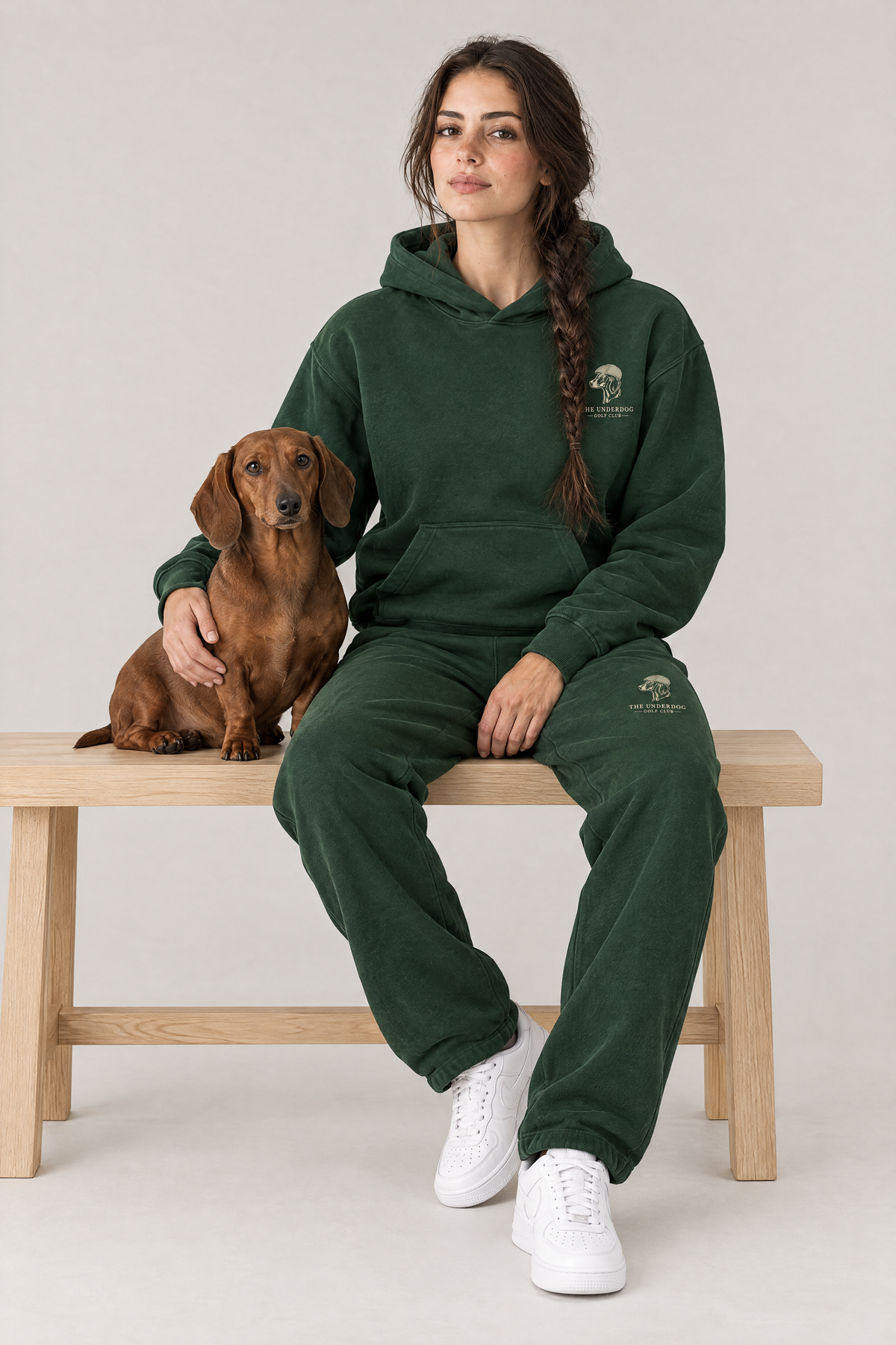 Woman sitting on a wooden bench with a red dachshund in The Long Drive dachshund golf hoodie and sweatpants in Blackish Green. Color: Blackish Green
