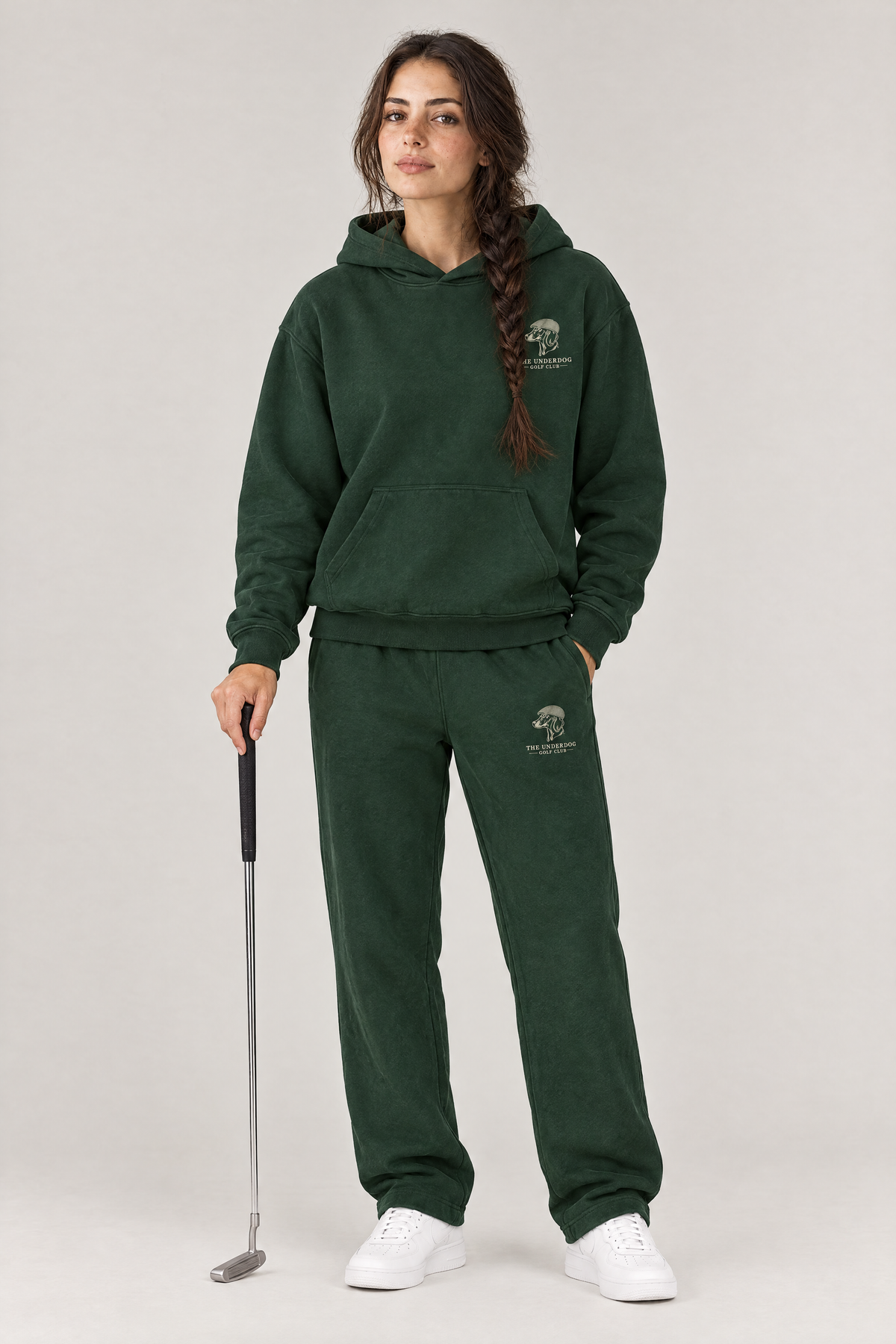 Woman holding a putter in The Long Drive dachshund golf hoodie and matching Underdog Golf Club sweatpants in Blackish Green, front view. Color: Blackish Green