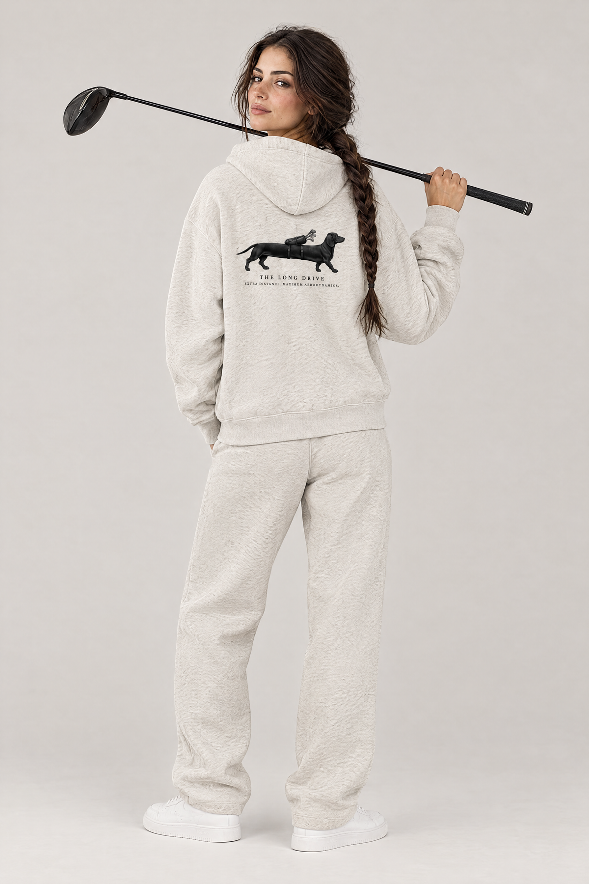 Woman with a driver over her shoulder, back view of The Long Drive dachshund golf hoodie in Oat Gray with the large back print. Color: Oat Gray