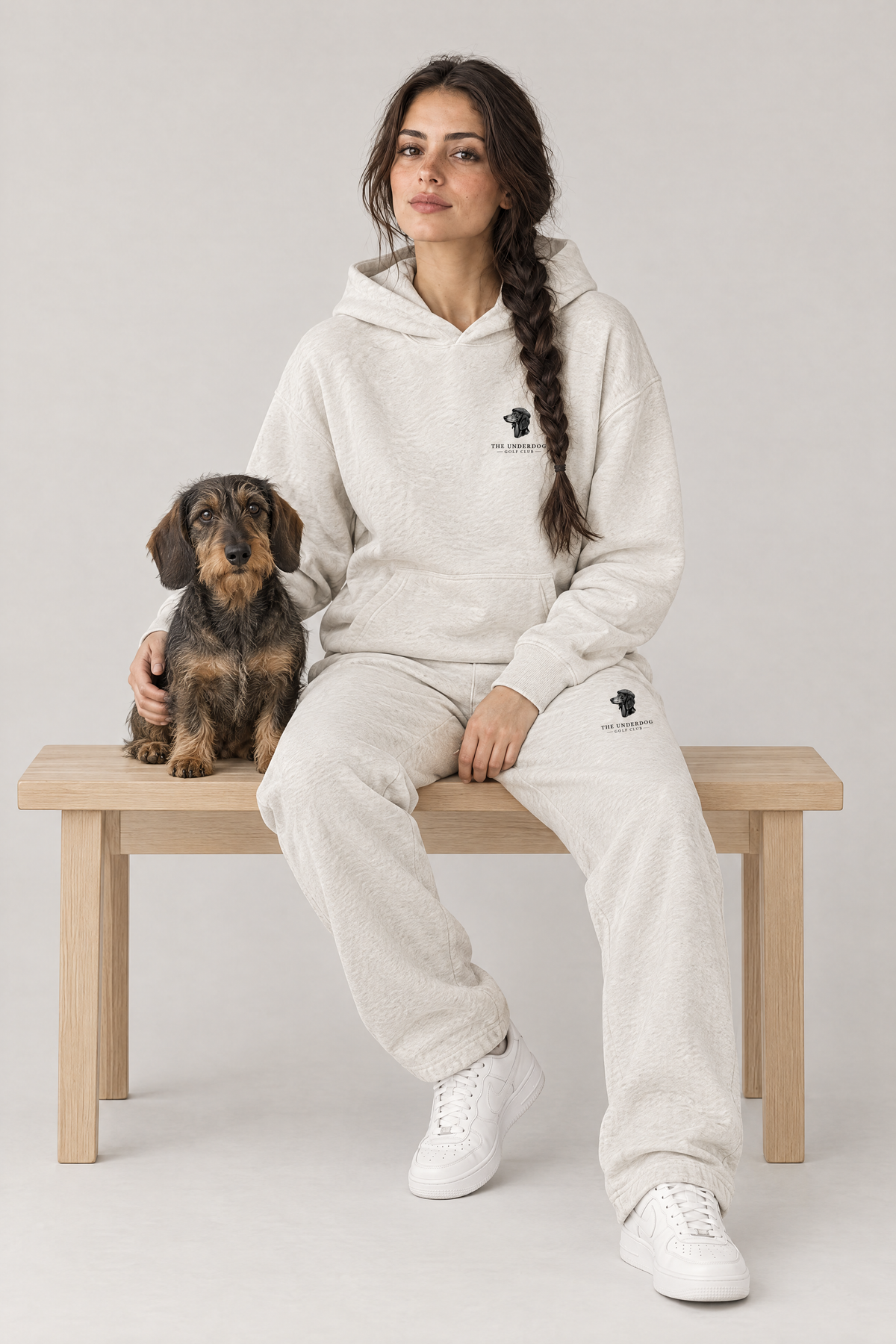 Woman sitting on a wooden bench with a wire-haired dachshund in The Long Drive dachshund golf hoodie and sweatpants in Oat Gray. Color: Oat Gray