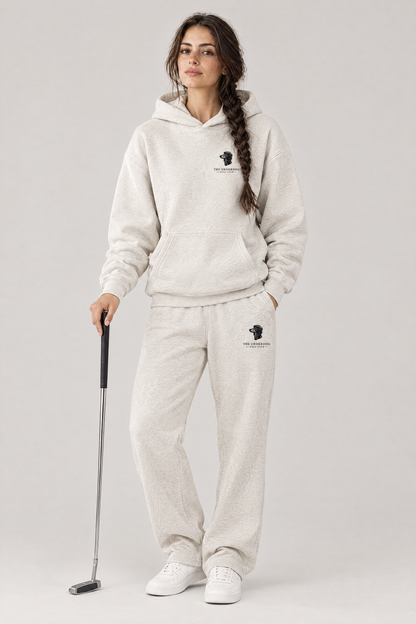 Woman holding a putter in The Long Drive dachshund golf hoodie and matching Underdog Golf Club sweatpants in Oat Gray, front view. Color: Oat Gray