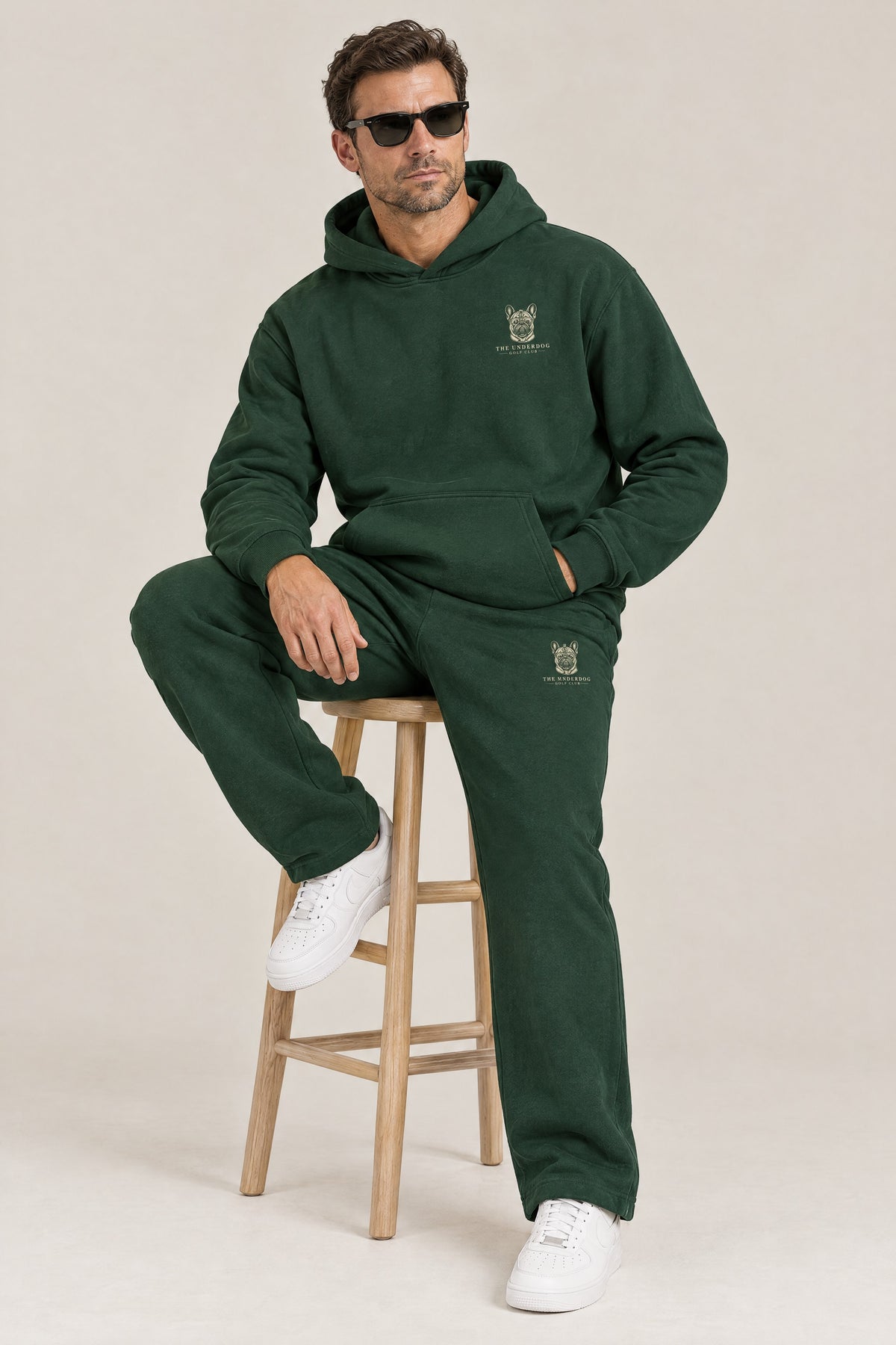 Man seated in The Marshal French Bulldog golf hoodie and sweatpants in Blackish Green. Color: Blackish Green