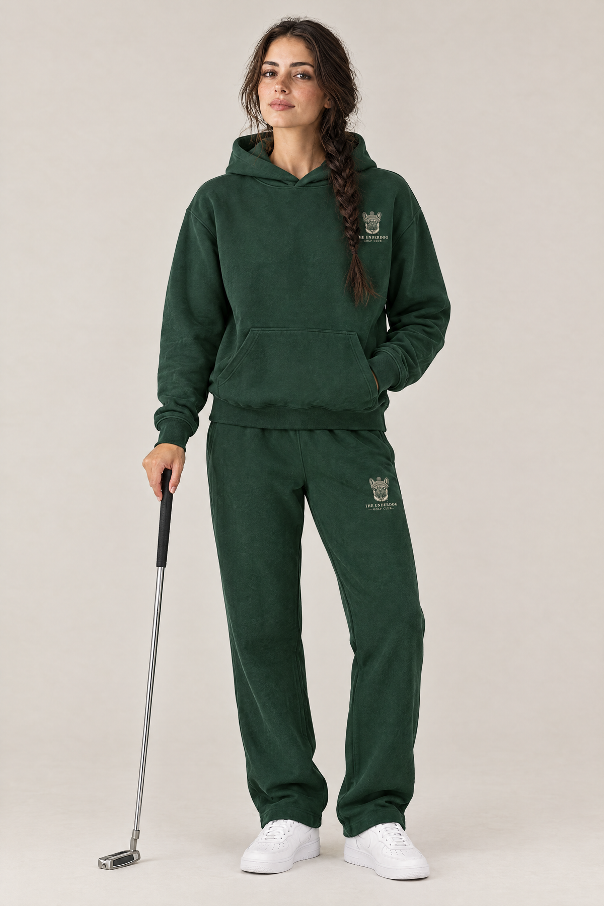 Woman holding a putter in The Marshal French Bulldog golf hoodie and matching Underdog Golf Club sweatpants in Blackish Green, front view. Color: Blackish Green
