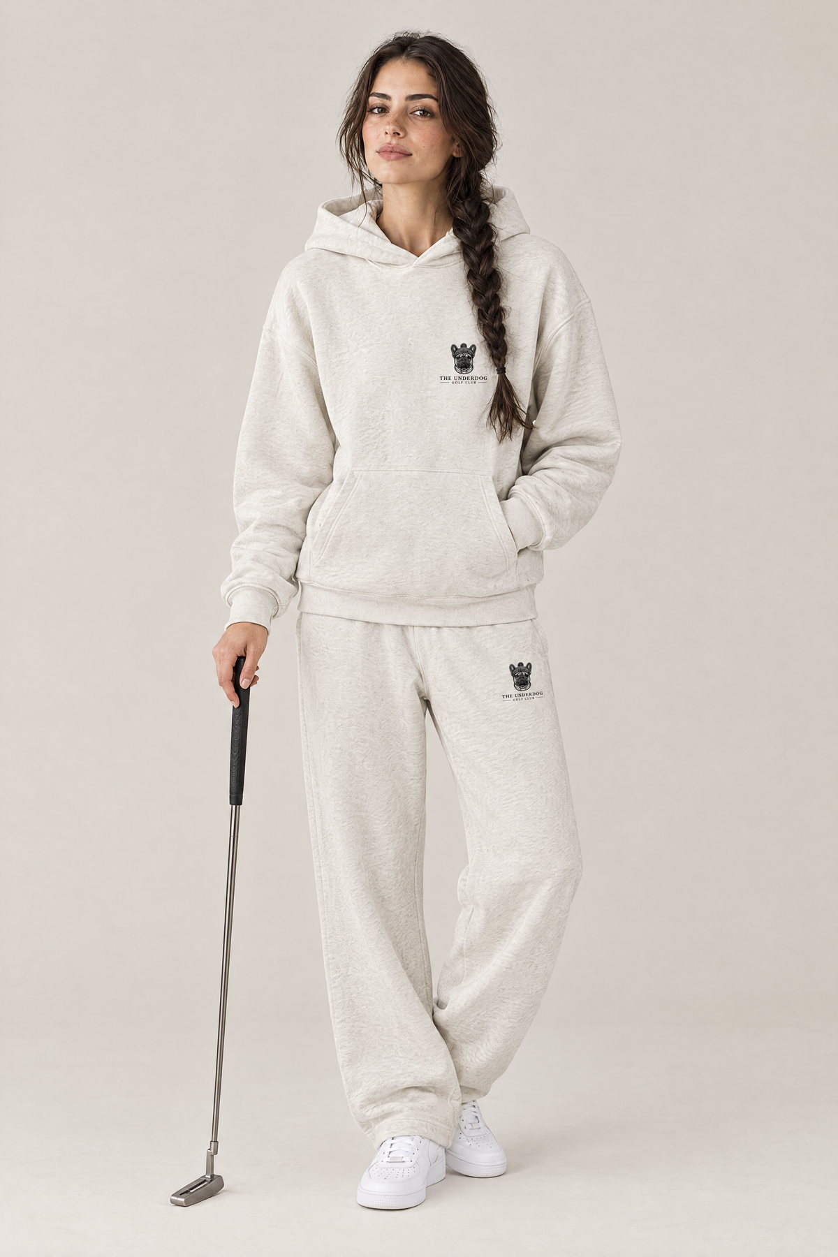 Woman holding a putter in The Marshal French Bulldog golf hoodie and matching Underdog Golf Club sweatpants in Oat Gray, front view. Color: Oat Gray
