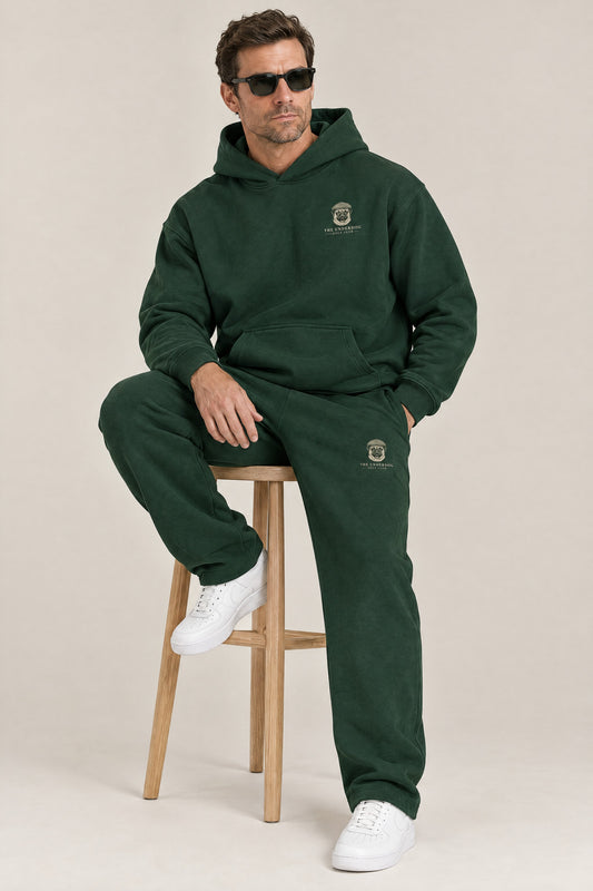 Man seated in The Heavy Hitter pug golf hoodie and sweatpants in Blackish Green. Color: Blackish Green