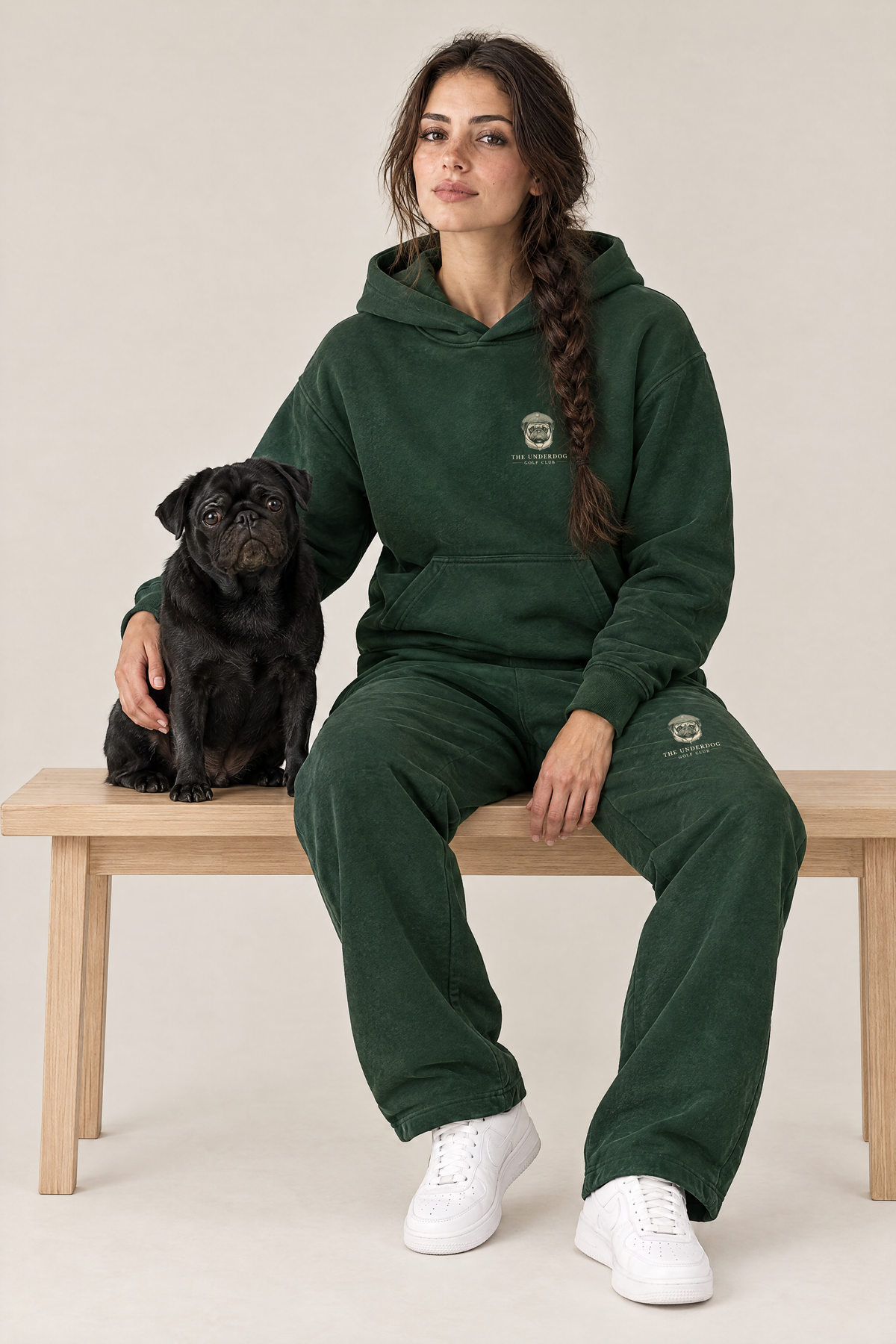 Woman sitting on a wooden bench with a black pug in The Heavy Hitter pug golf hoodie and sweatpants in Blackish Green. Color: Blackish Green