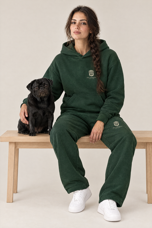 Woman sitting on a wooden bench with a black pug in The Heavy Hitter pug golf hoodie and sweatpants in Blackish Green. Color: Blackish Green