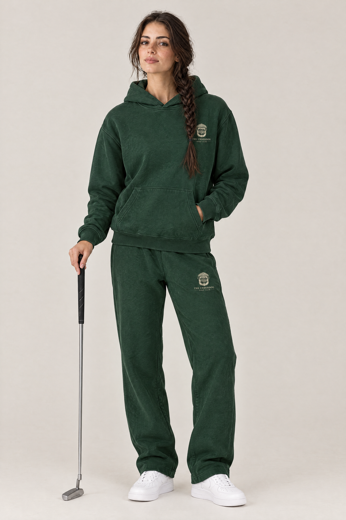 Woman holding a putter in The Heavy Hitter pug golf hoodie and matching Underdog Golf Club sweatpants in Blackish Green, front view. Color: Blackish Green