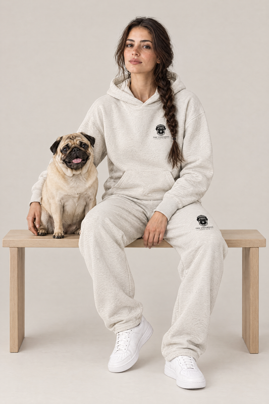 Woman sitting on a wooden bench with a fawn pug in The Heavy Hitter pug golf hoodie and sweatpants in Oat Gray. Color: Oat Gray