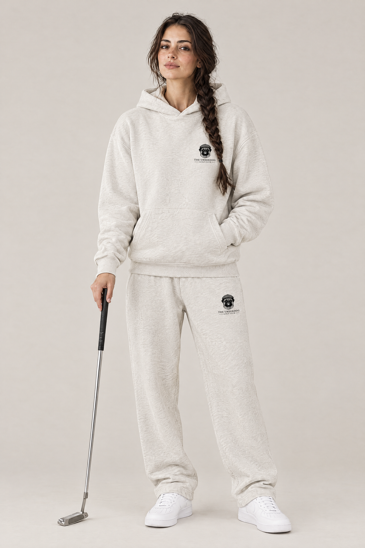 Woman holding a putter in The Heavy Hitter pug golf hoodie and matching Underdog Golf Club sweatpants in Oat Gray, front view. Color: Oat Gray