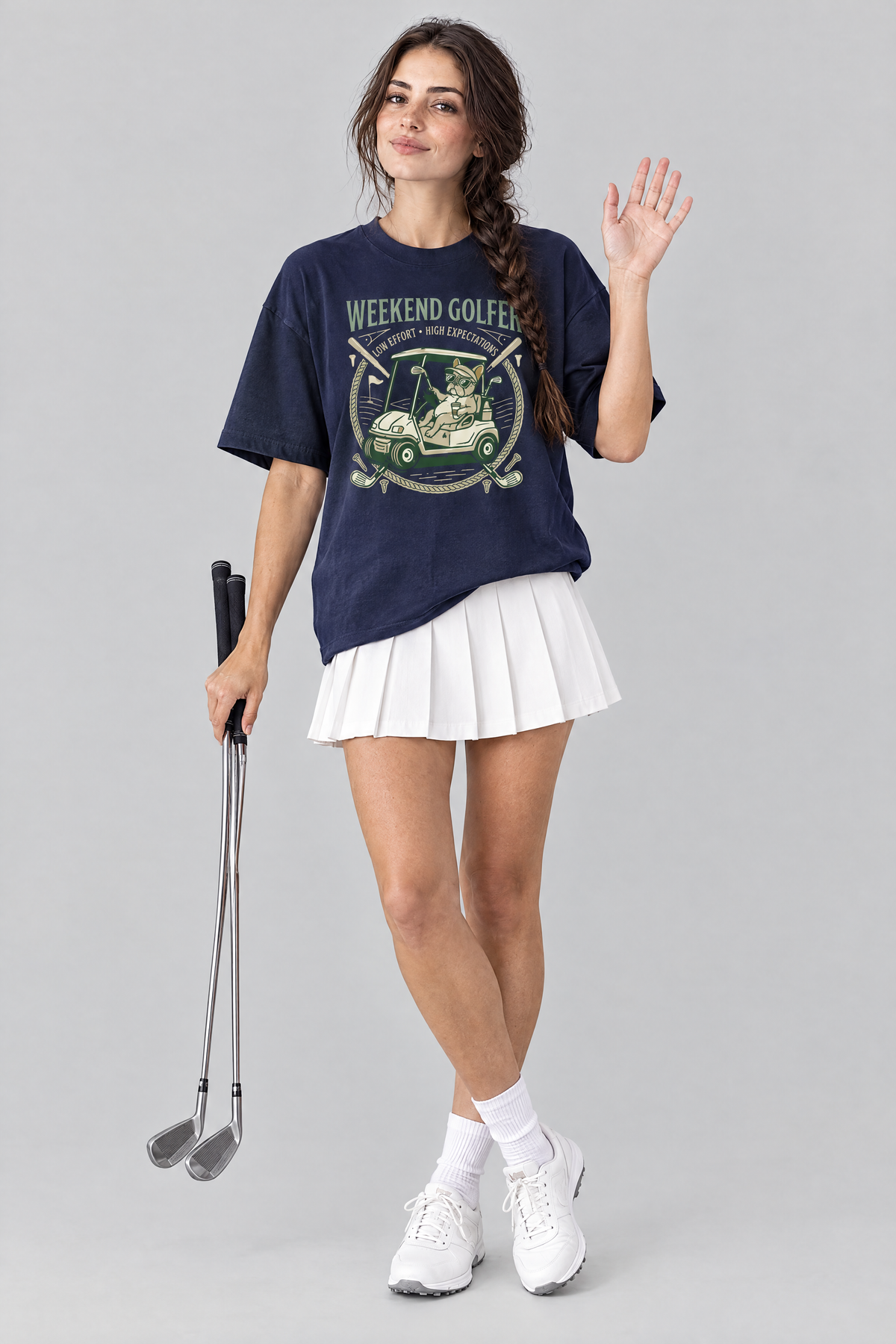 Woman waving and holding golf clubs in the navy Weekend Golfer French Bulldog Golf Tee and a white pleated skirt. Color: Navy
